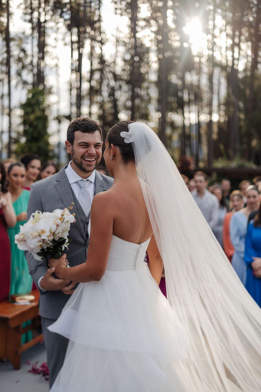 A wedding celebration outdoors with sunlight filtering through trees as couple shares joyful moment surrounded by guests.