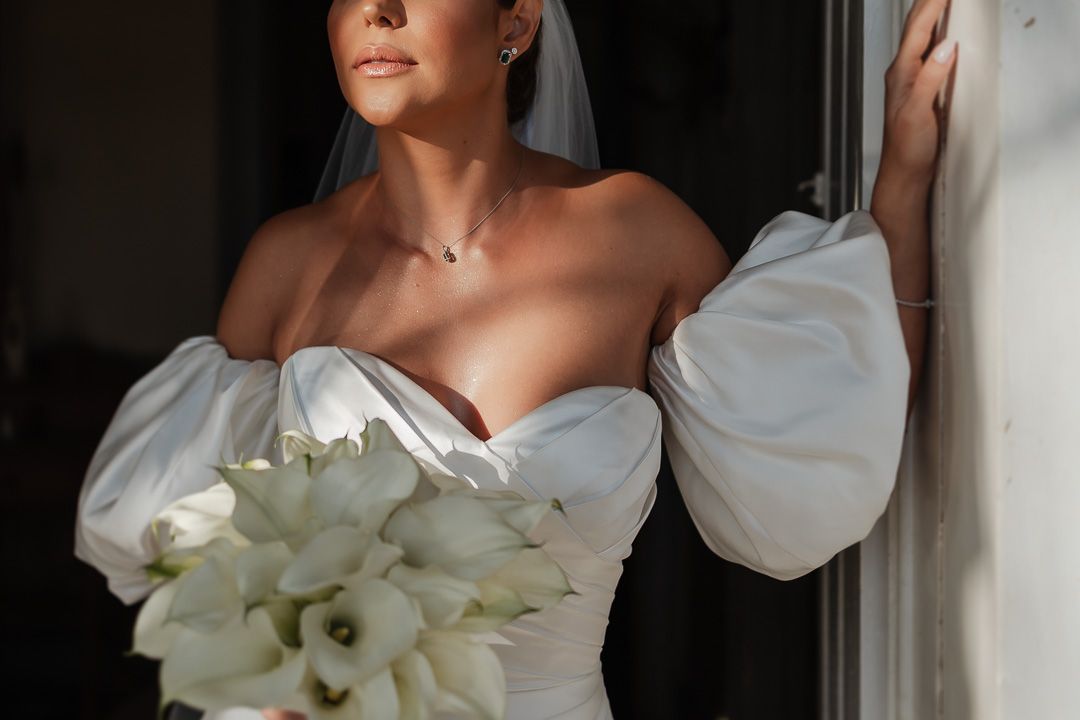 Elegant bride in white off-shoulder wedding dress holds bouquet of white calla lilies by window.