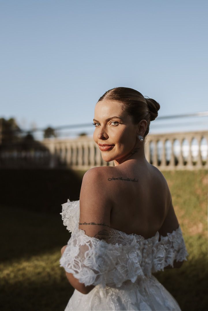 A person wearing an off-shoulder white lace wedding dress poses outdoors at sunset next to a balustrade.