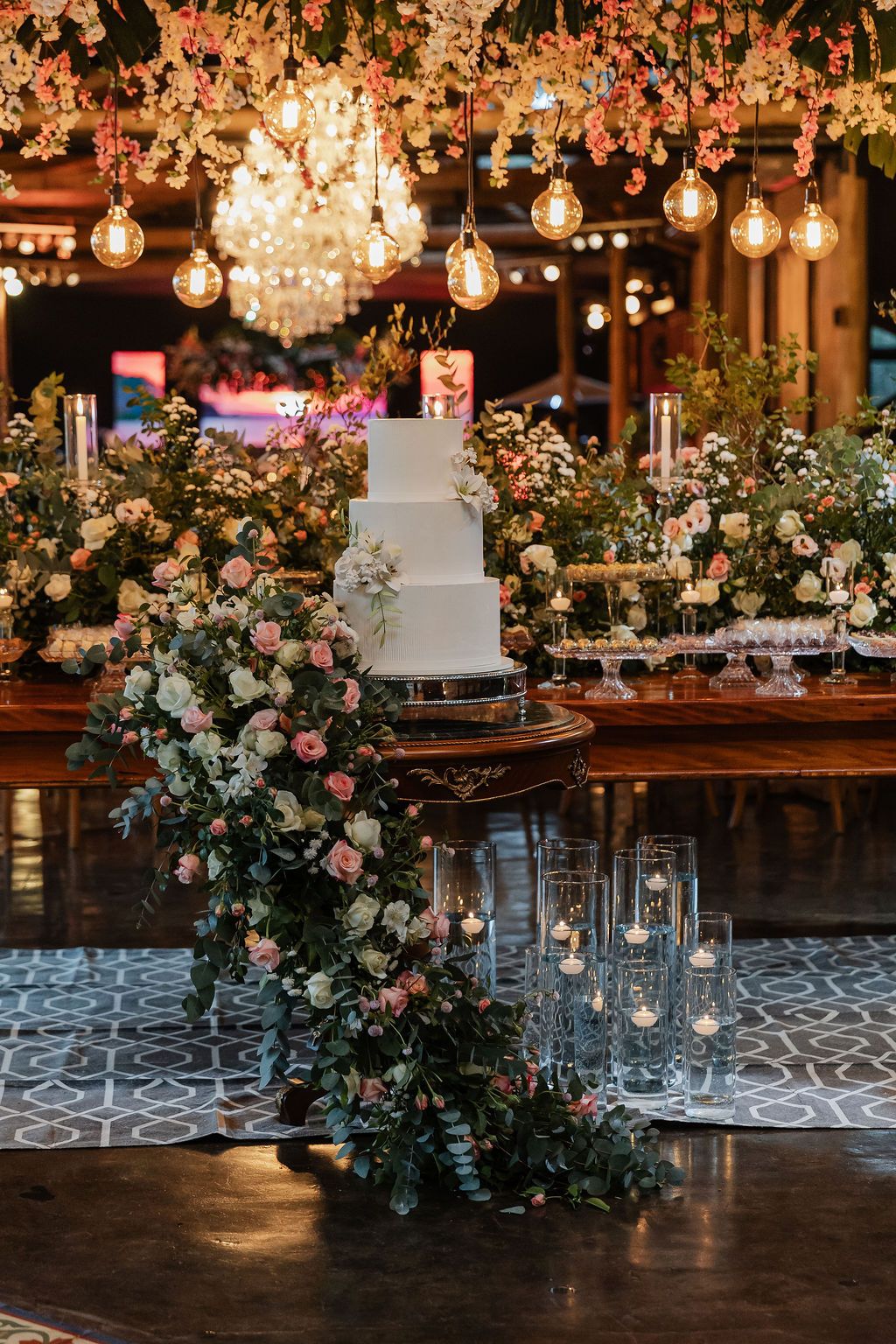 Elegant white wedding cake surrounded by floral arrangements on wooden table with hanging Edison bulbs creating warm ambiance.