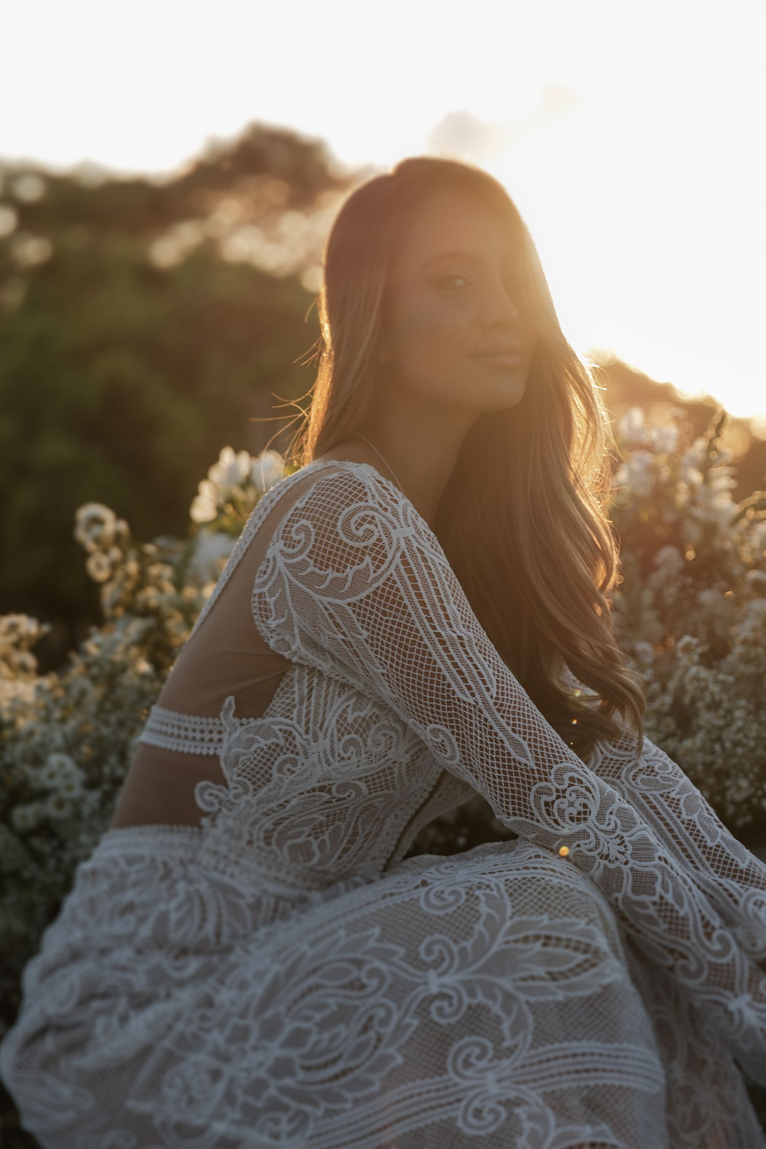 A delicate white lace wedding dress is illuminated by golden sunlight against a natural outdoor backdrop.