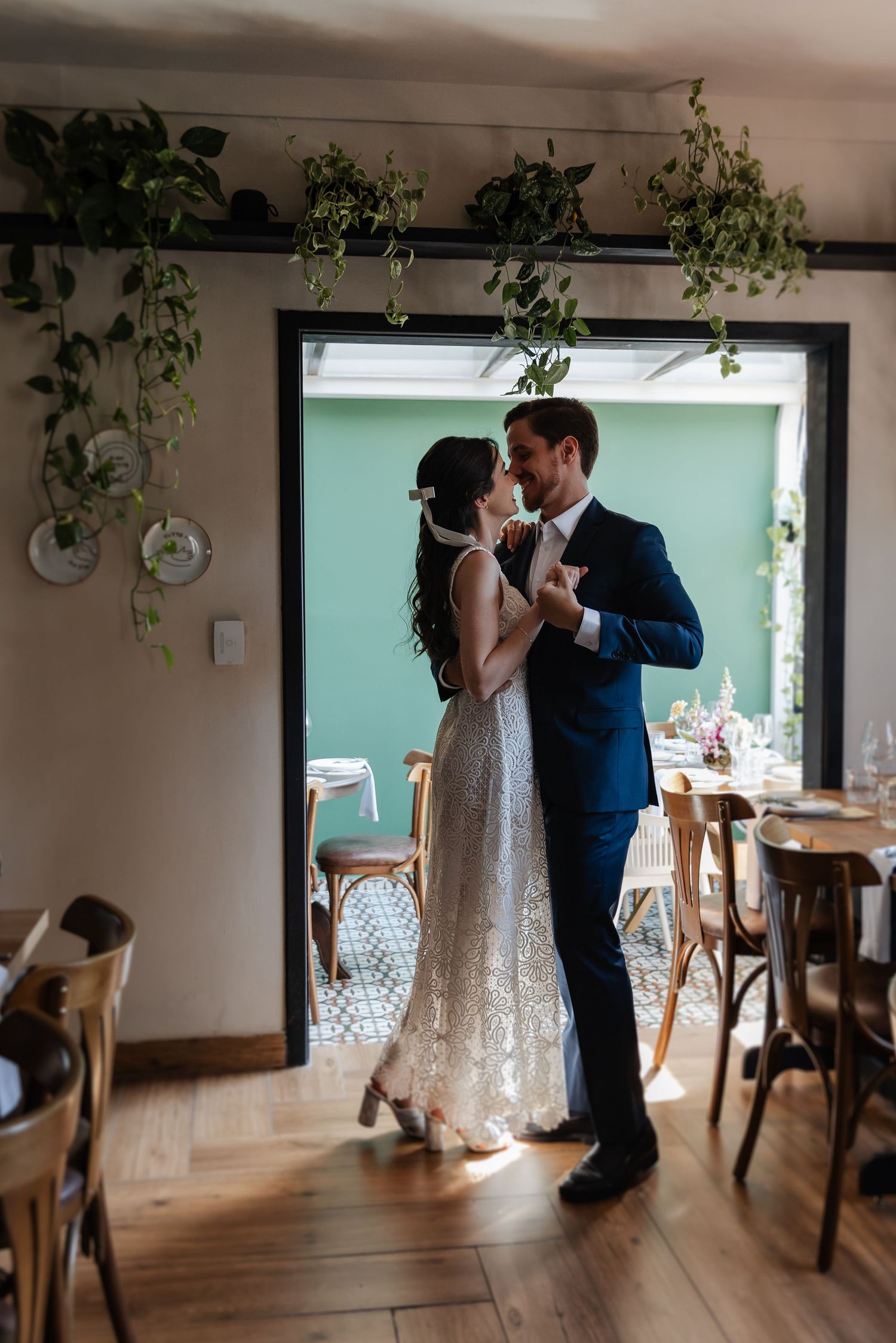 A romantic wedding couple dances together in a light-filled room with wooden floors and hanging plants.