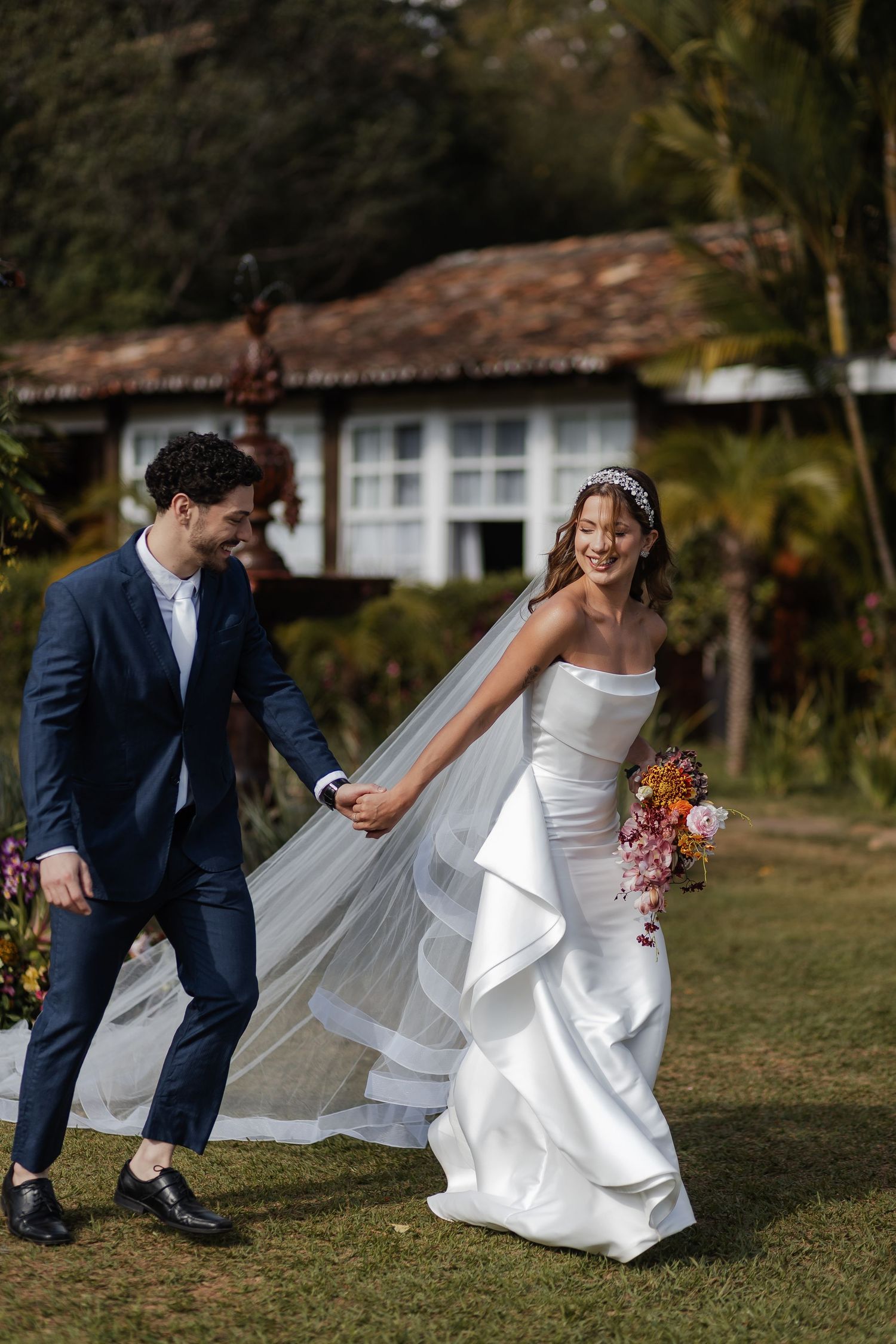 A couple shares a playful moment outdoors at their tropical wedding, with a rustic house and palm trees in the background.