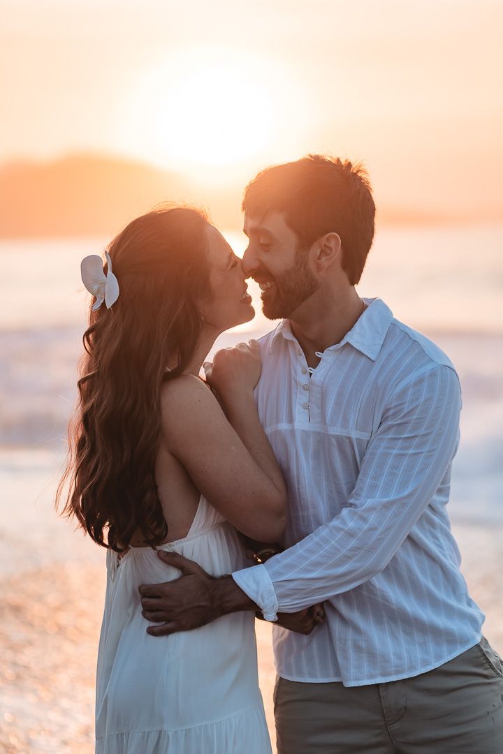 A couple shares an intimate moment at sunset on the beach, embracing in white summer outfits against the golden sky.