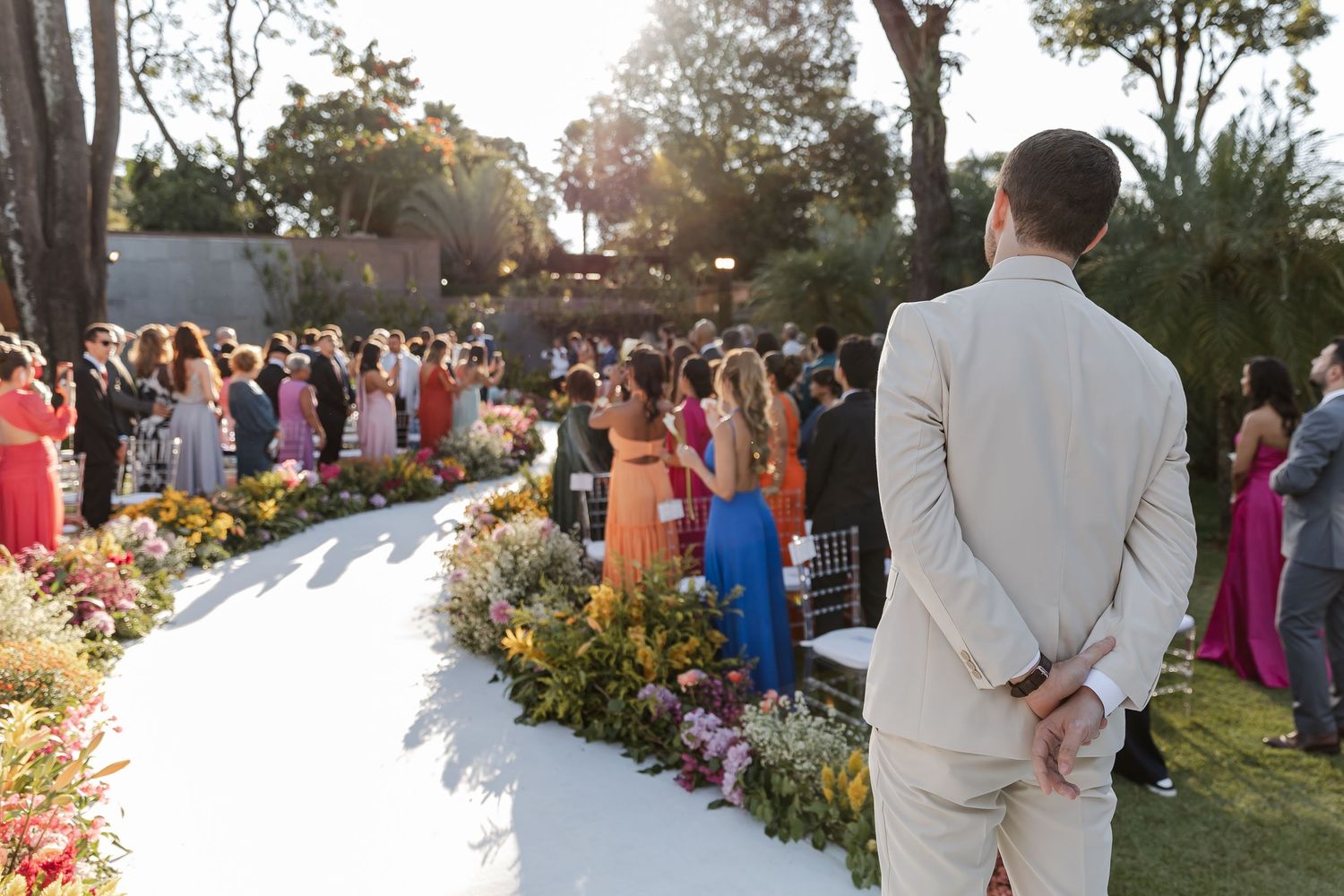 Beautiful outdoor wedding ceremony at sunset with guests in colorful formal attire gathered along a flower-lined aisle.