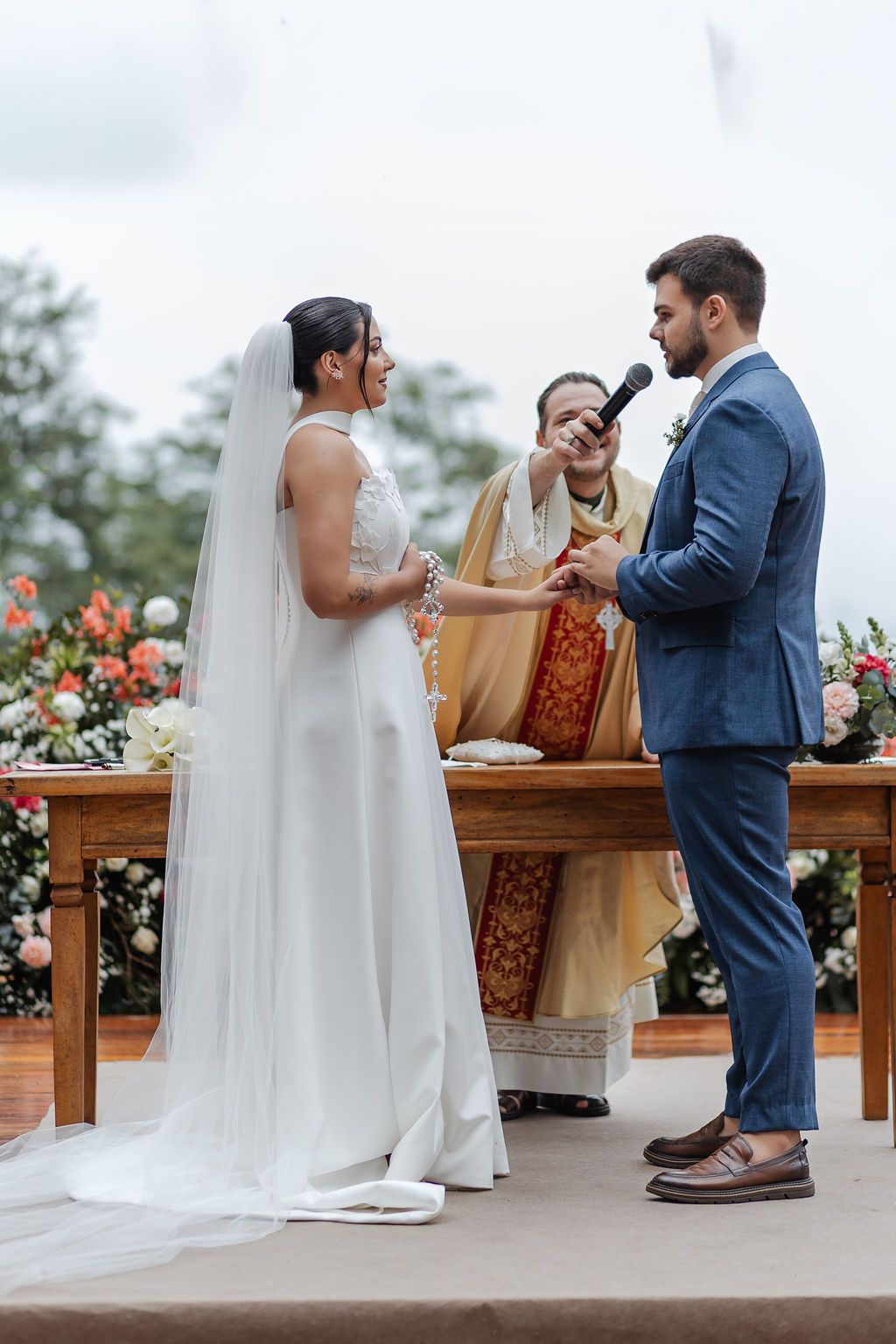 A wedding ceremony takes place outdoors with a couple exchanging vows in front of wooden benches and floral arrangements.