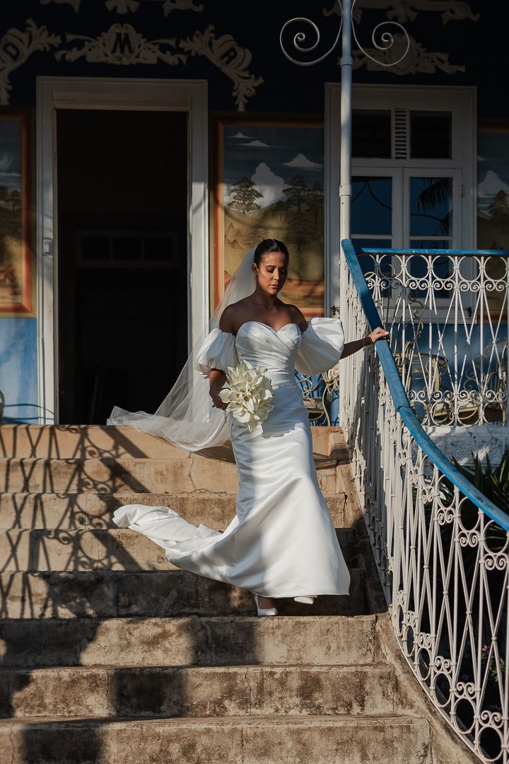 A bride in an elegant white gown and veil stands on stone steps with ornate wrought iron railings at a historic venue.