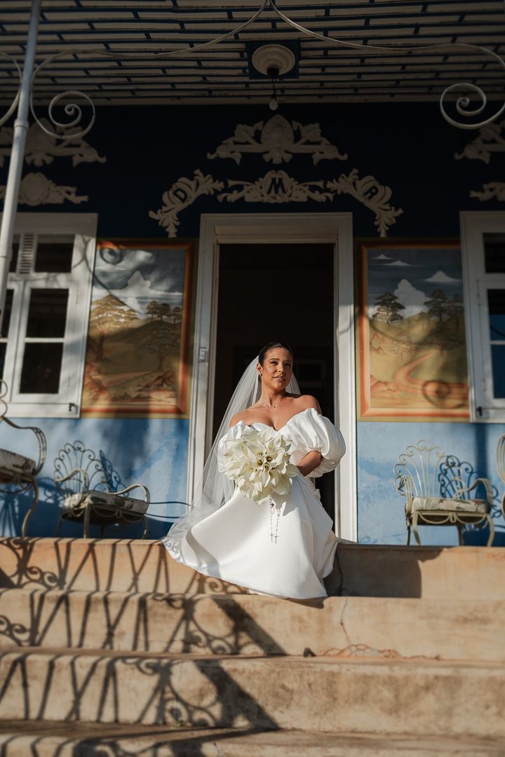 Bride in white off-shoulder gown poses on ornate porch with painted murals and decorative ironwork.
