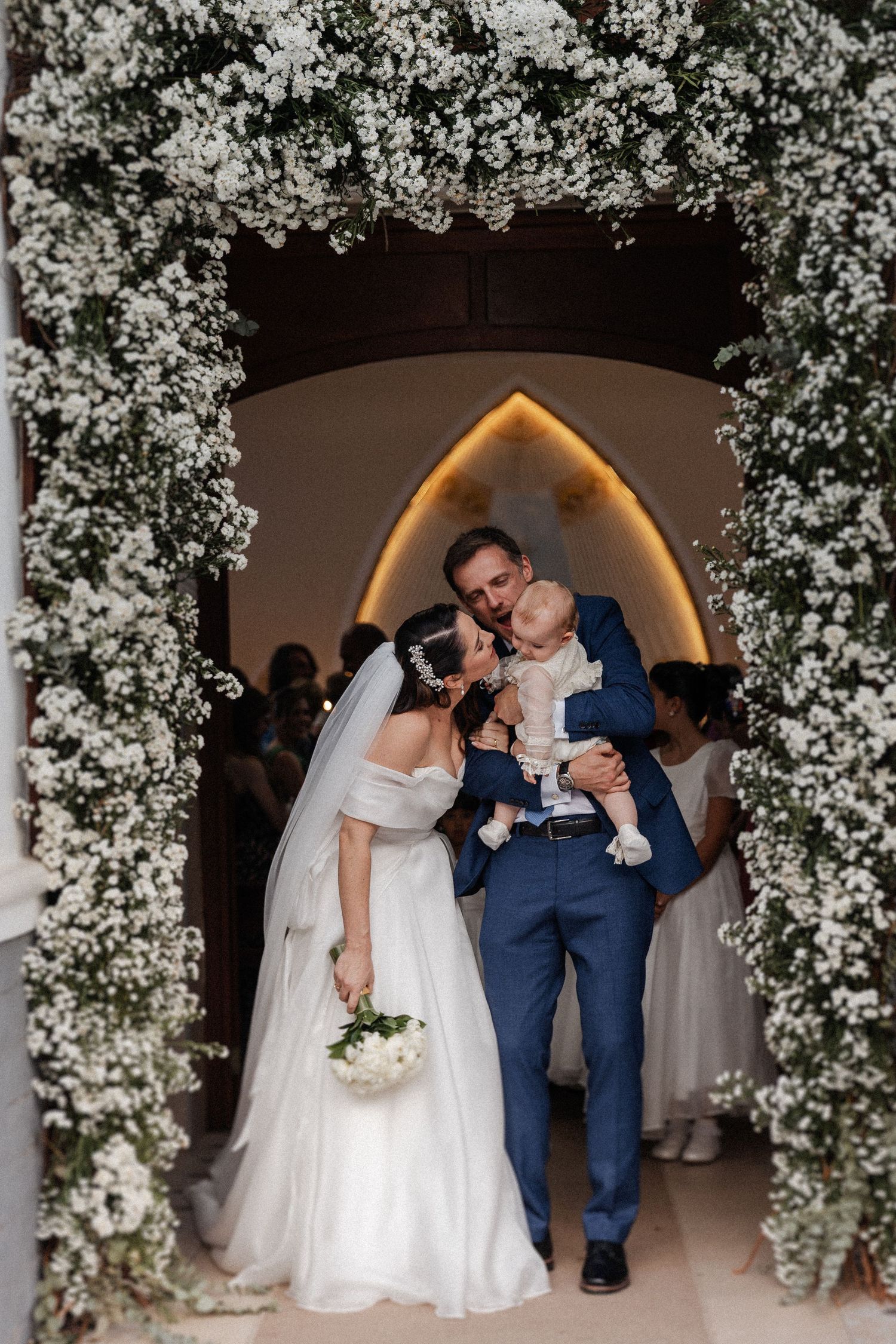 A romantic wedding couple shares a kiss under an archway decorated with cascading white flowers.