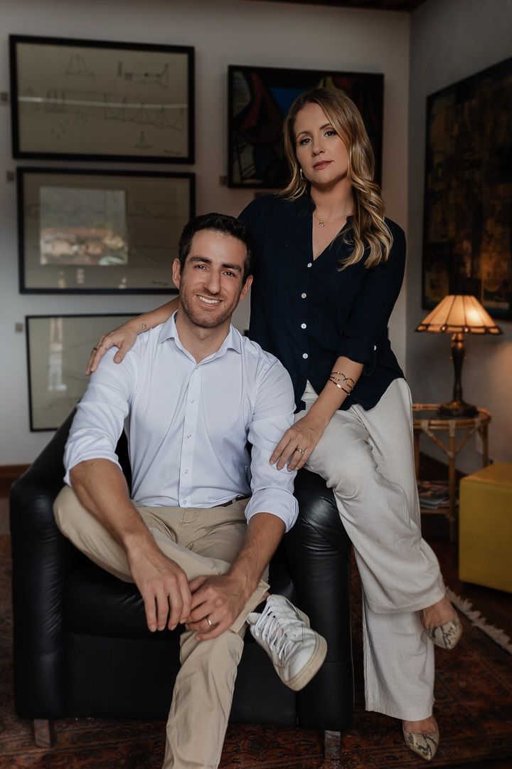 A couple poses together in a cozy living room setting with vintage decor, dark leather furniture and framed artwork.