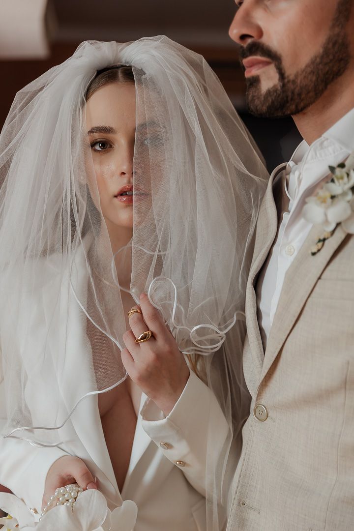 A bride in a white veil and gown stands close to a bearded person wearing a light colored suit at their wedding.