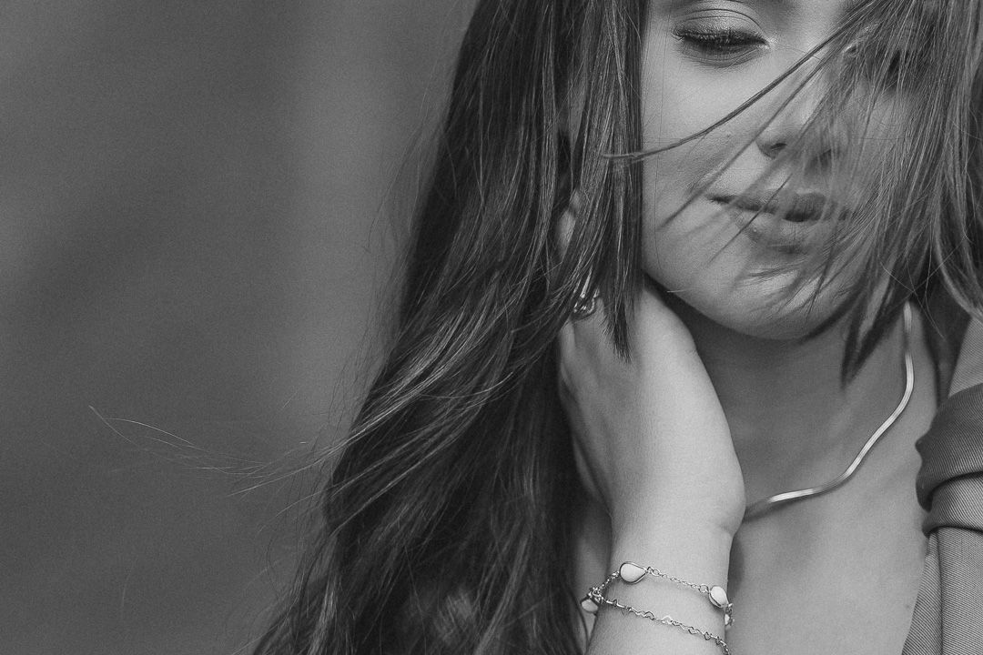 Artistic black and white portrait shot with long windswept hair and delicate jewelry details.