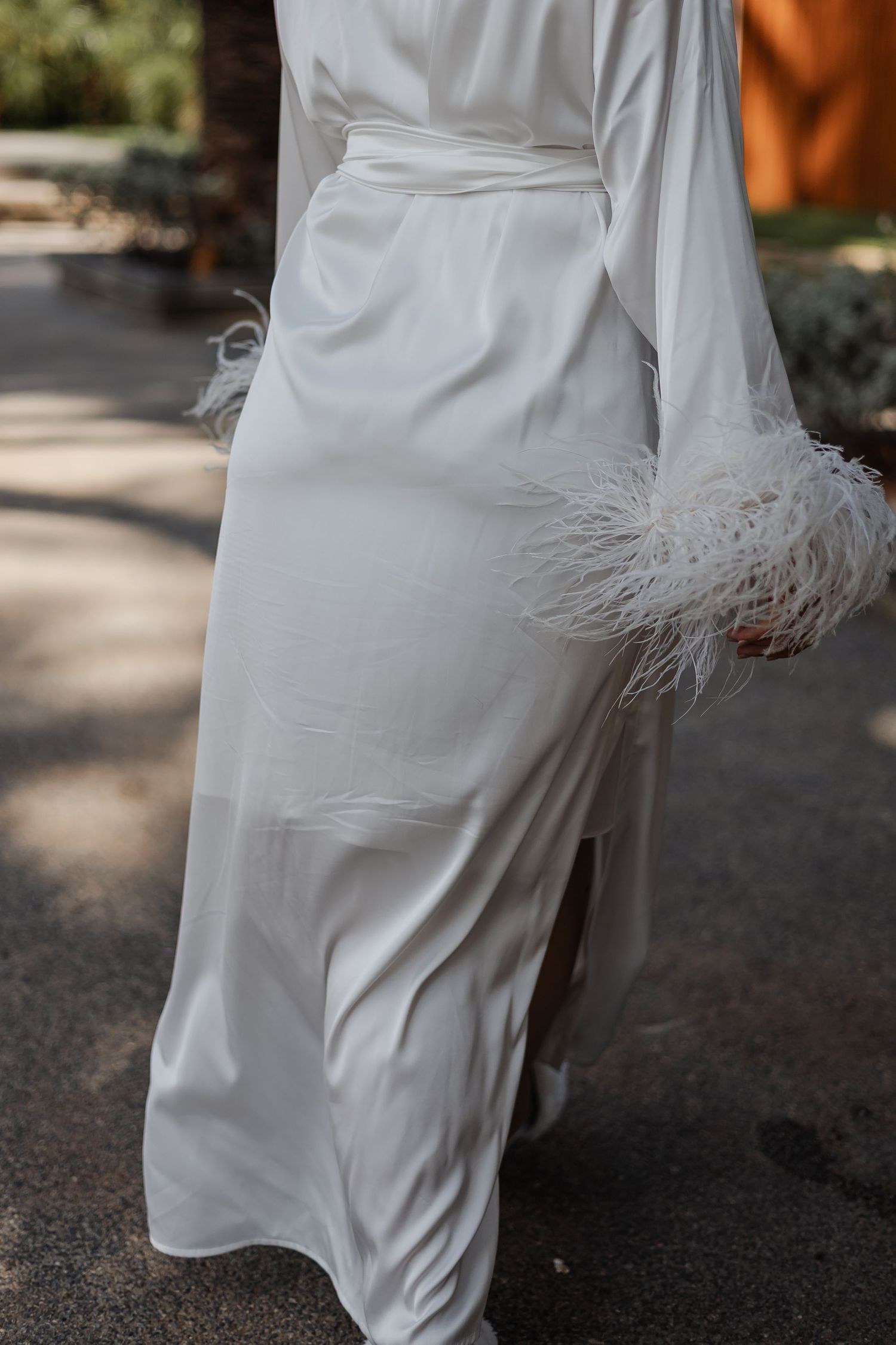 Elegant white satin wedding dress with feather trim sleeve detail shown in outdoor garden setting.