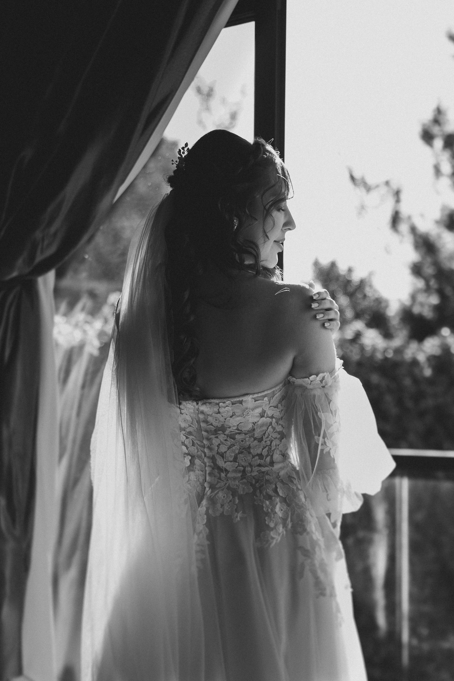 A bride in a lace wedding dress and veil stands by a window in black and white silhouette.