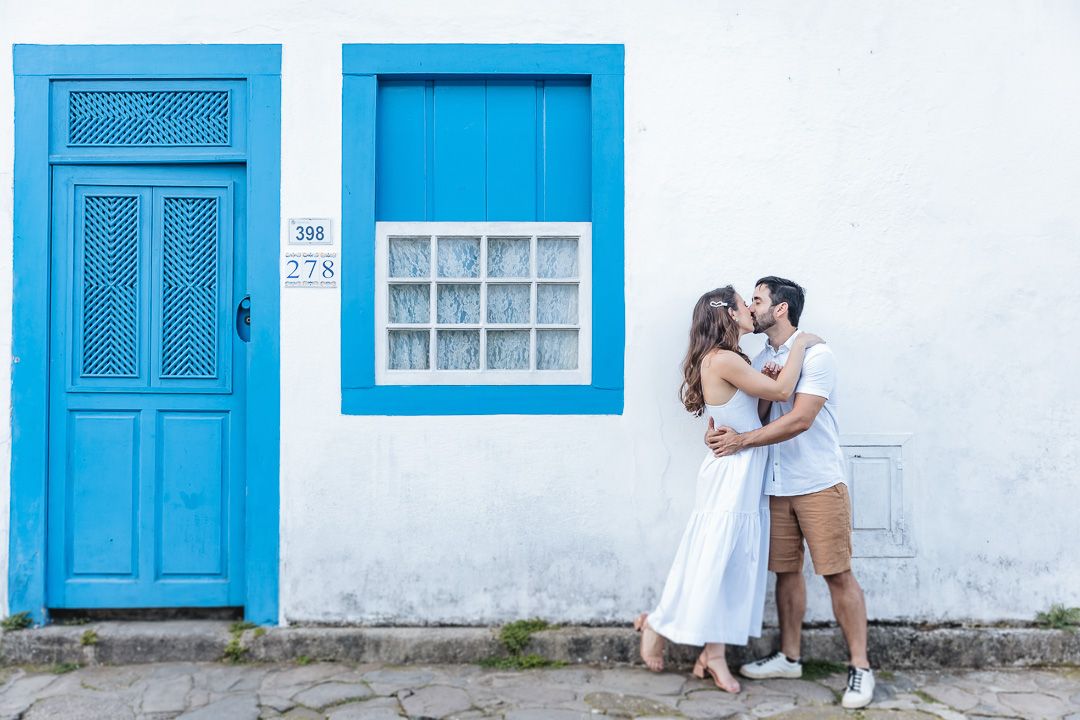 A couple embraces by a wall with bright blue door and window frames, creating a romantic Mediterranean-style scene.