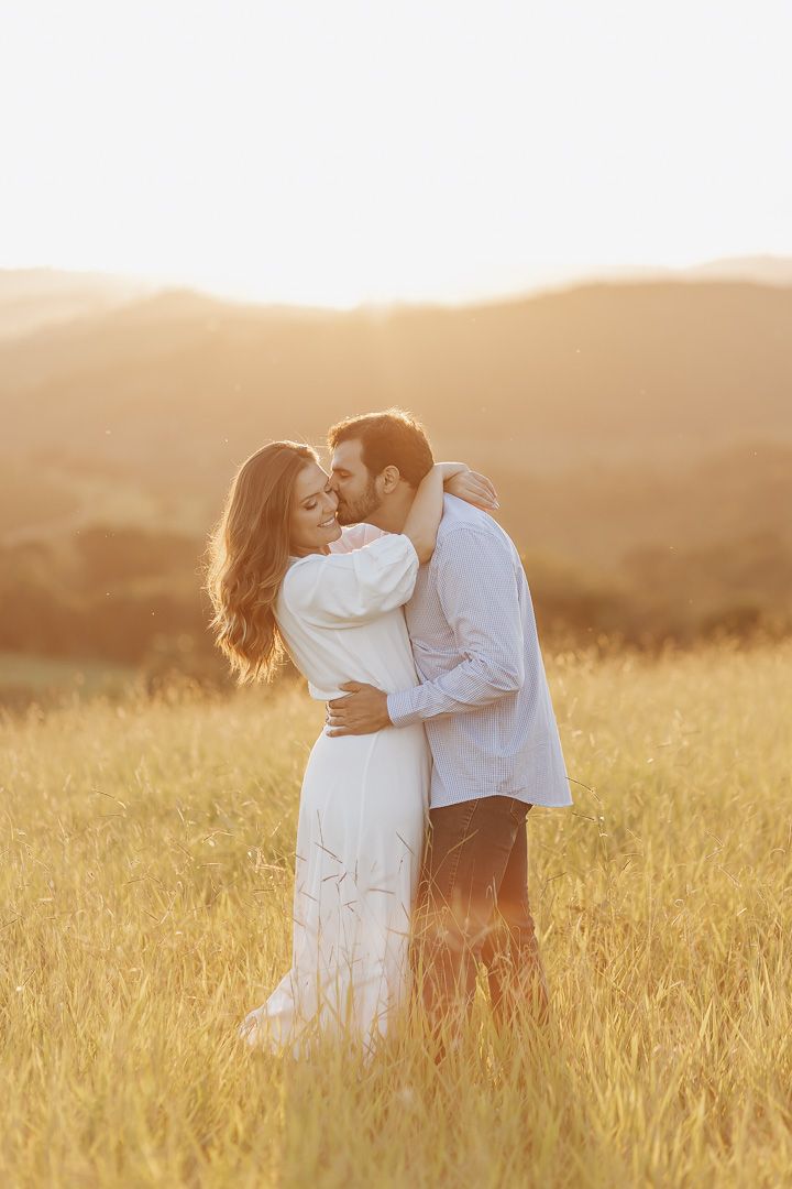 Romantic couple embraces in golden sunlit field during sunset with rolling hills in background creating dreamy atmosphere.