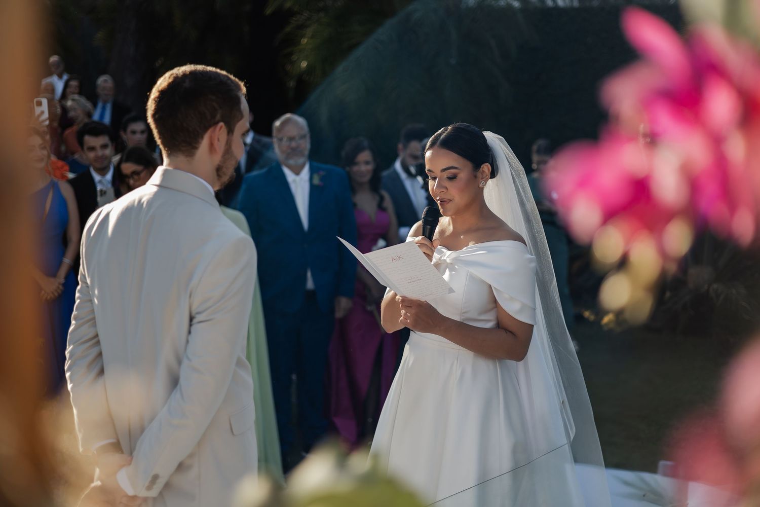 A bride reads from a paper during an outdoor wedding ceremony with pink flowers in the foreground.