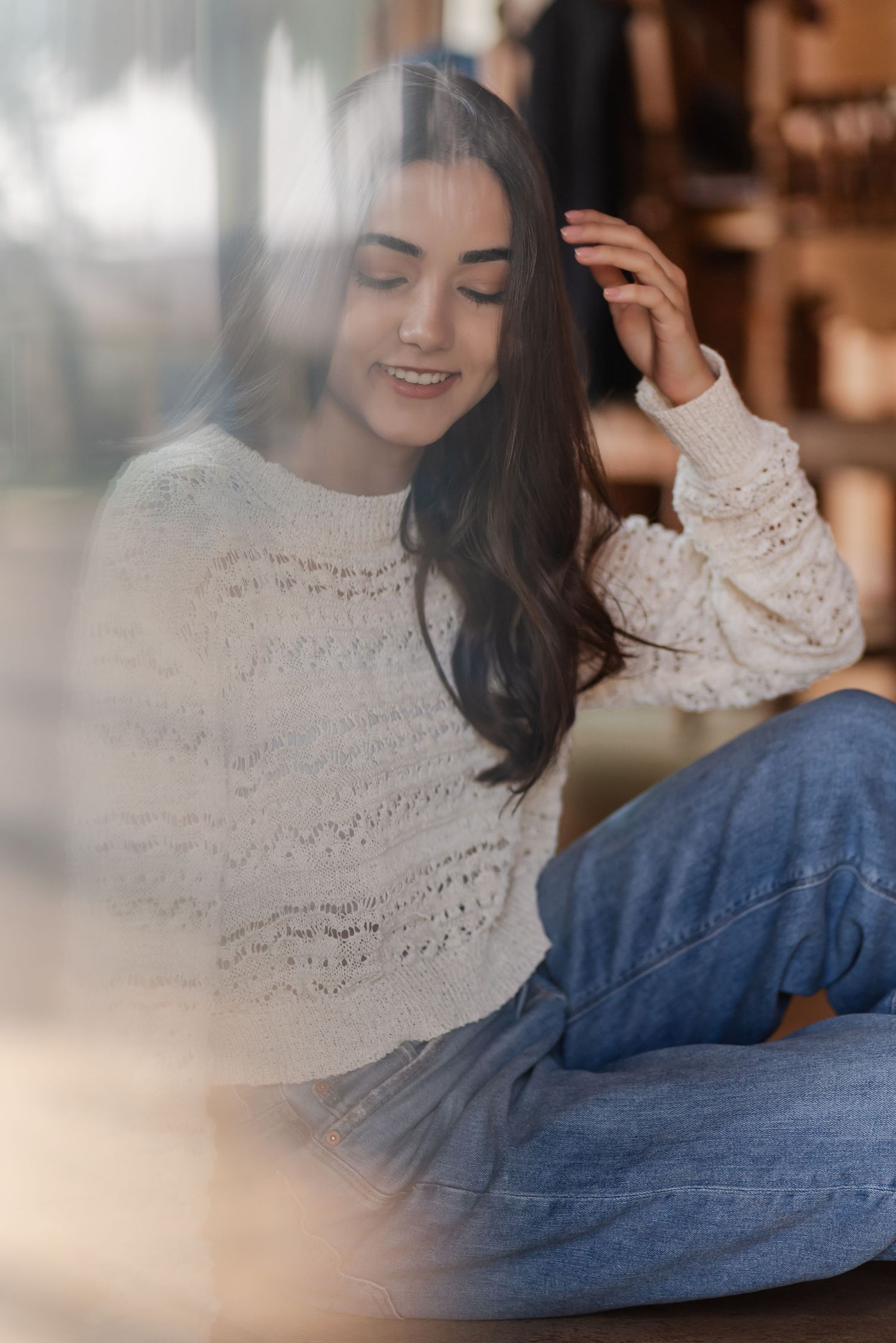 Portrait in dreamy lighting showing someone in a cream knit sweater and jeans relaxing with a soft smile and long dark hair.