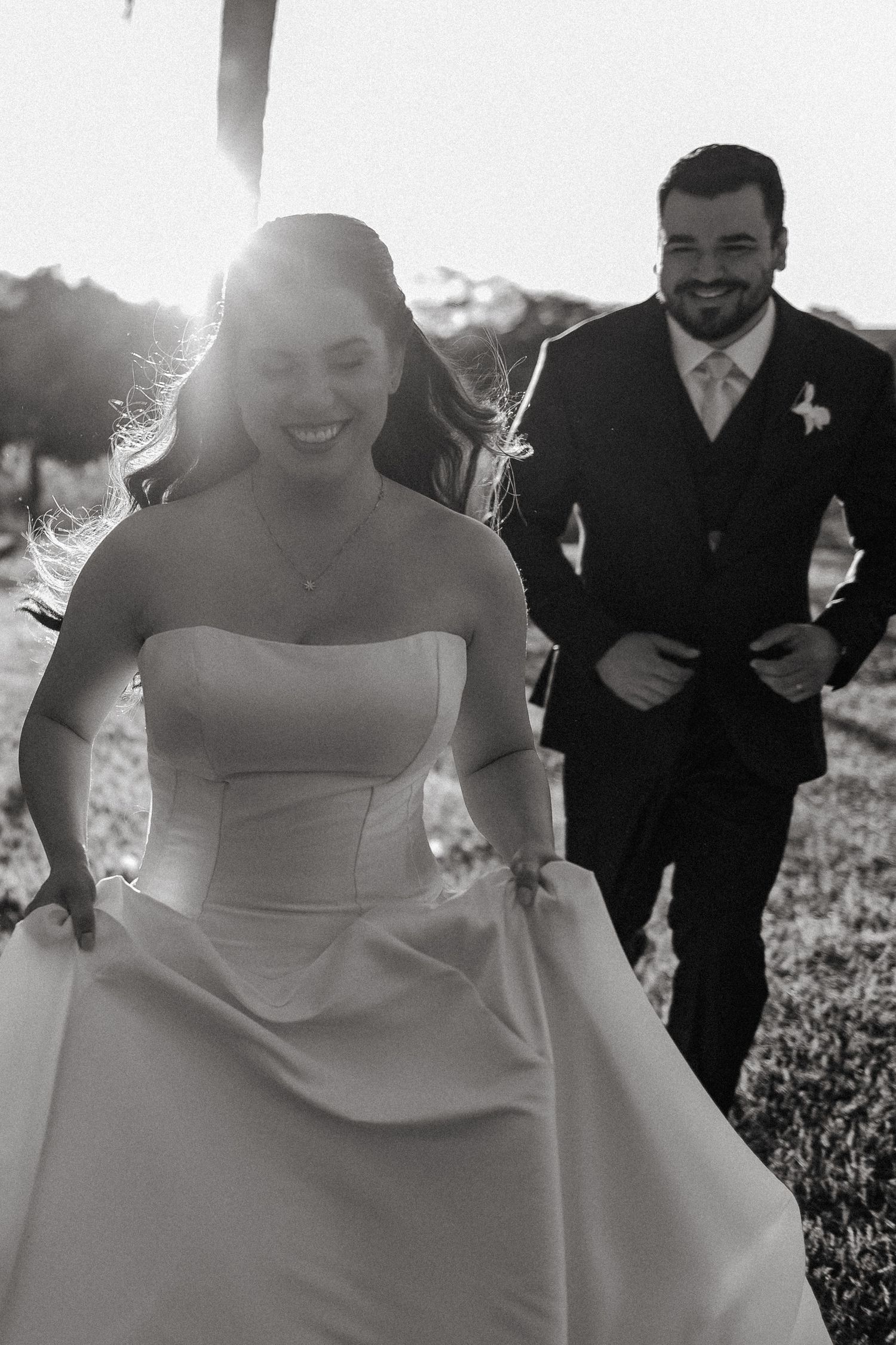 A joyful wedding scene shows a bride in a white strapless gown laughing and walking outdoors on a sunny day.