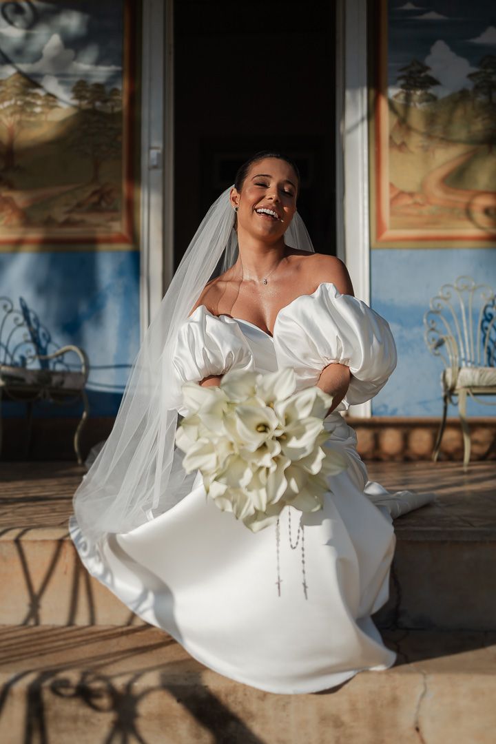 A bride in a stunning off-shoulder wedding gown holds a large white bouquet while posing outdoors in warm sunlight.