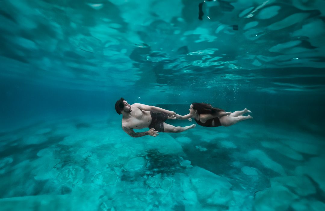 Two swimmers float peacefully in turquoise underwater scene with sunlight filtering through from above.