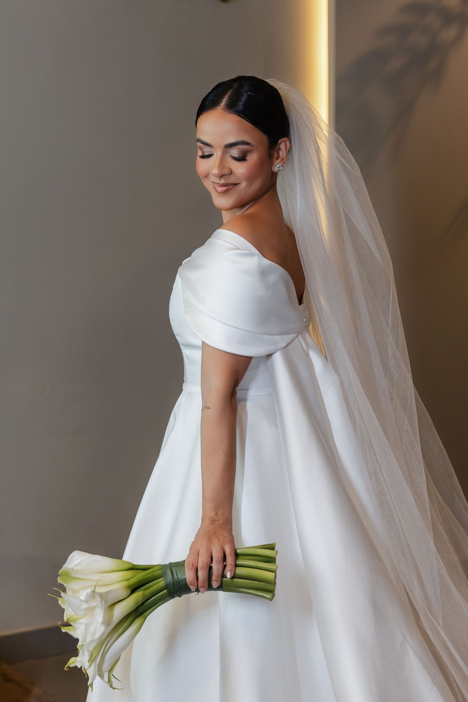 A bride in an elegant white off-shoulder wedding dress holds a classic white calla lily bouquet while wearing a flowing veil.
