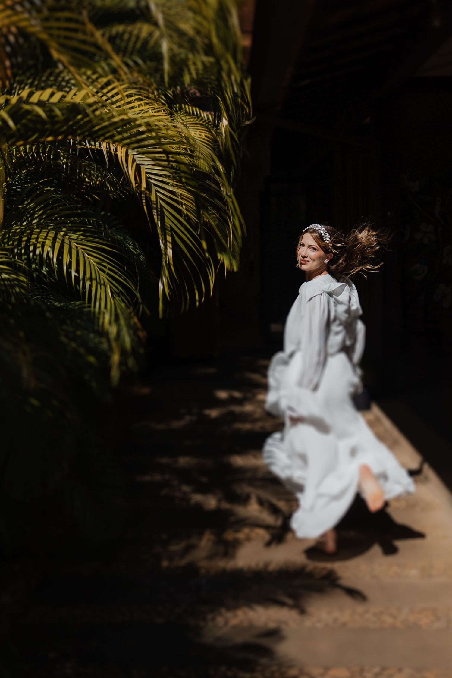 A flowing white dress billows in the breeze against tropical palm fronds casting dramatic shadows.