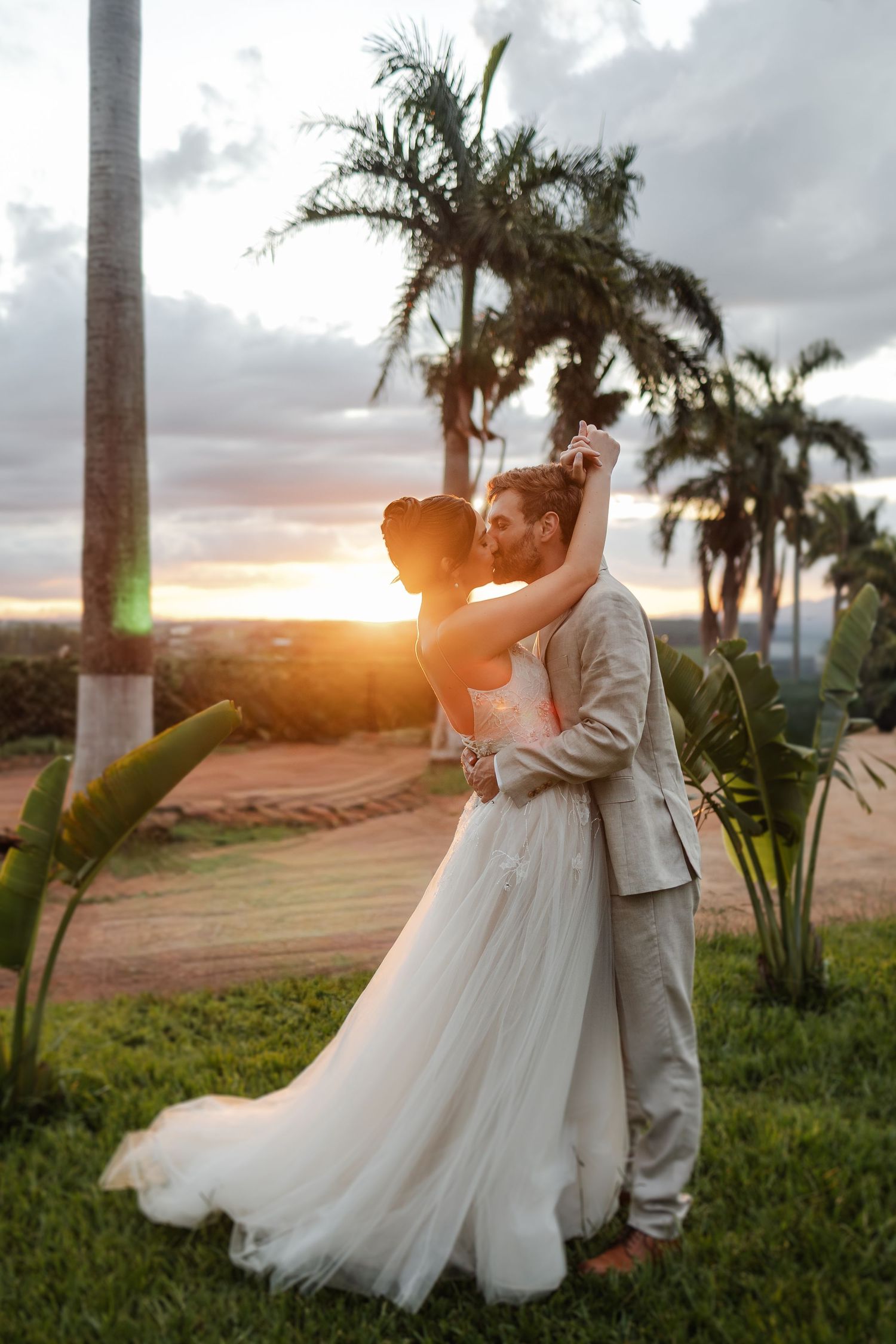 A romantic sunset wedding moment with palm trees silhouetted against a glowing orange sky in a tropical garden setting.