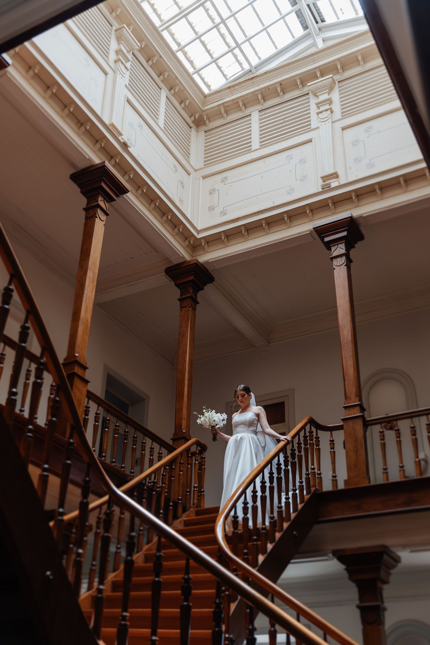 Elegant wooden staircase in historic building with skylight illuminating a figure in white dress descending the steps.