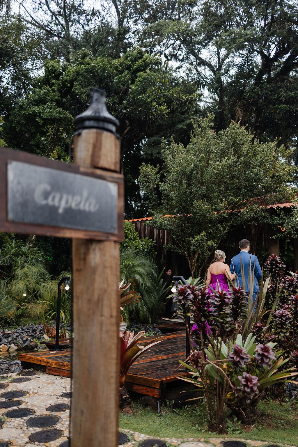 Inspire-se com o casamento ao aWooden signpost reading 'Capela' stands near a garden path with lush tropical plants and a decorative bridge.