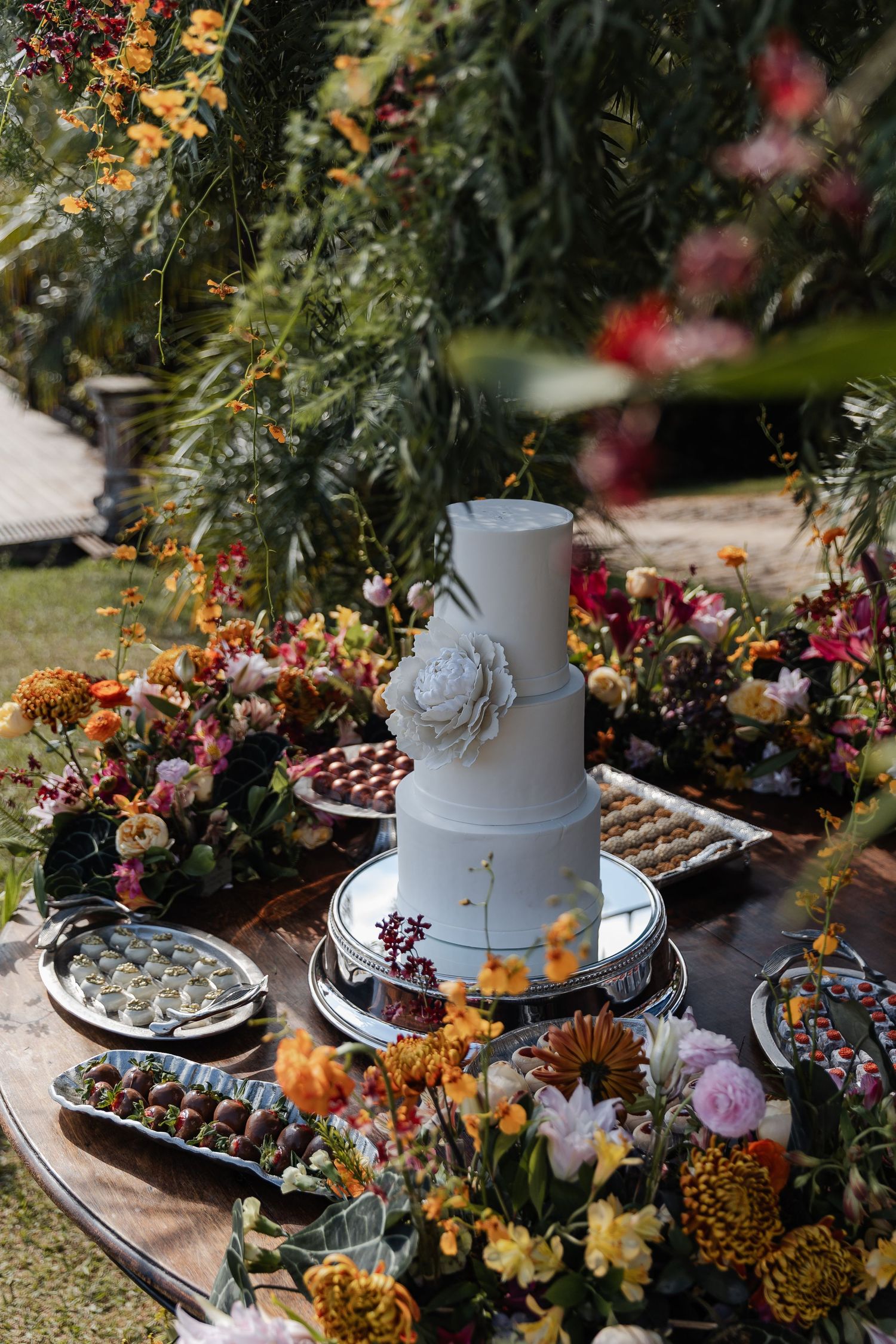 White tiered cake displayed on dessert table decorated with colorful autumn flowers and greenery at outdoor celebration.