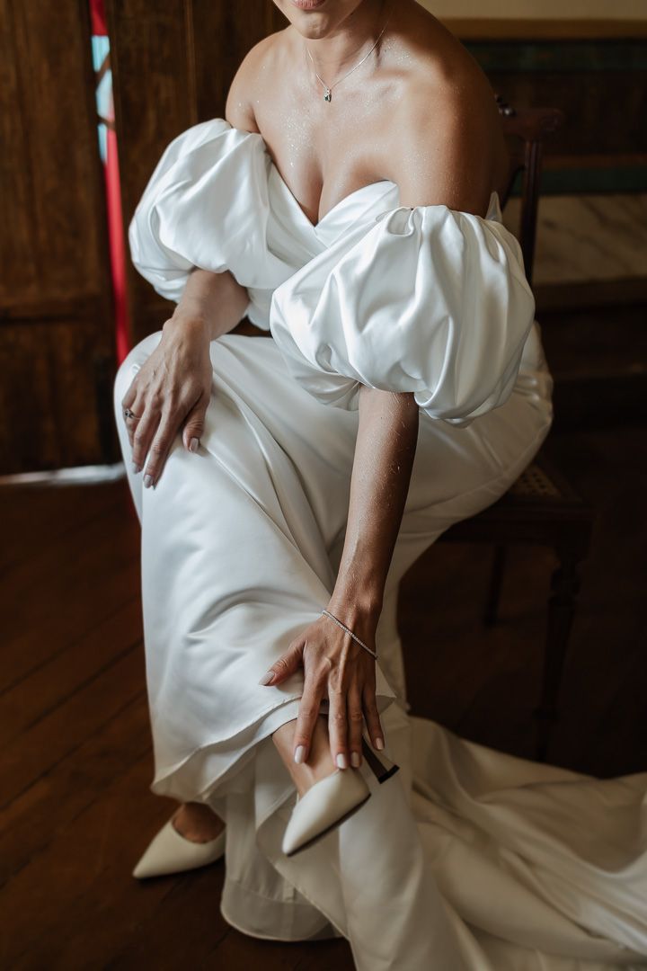 A white wedding dress with dramatic puff sleeves and off-shoulder neckline is styled with elegant pointed heels on wooden floor.