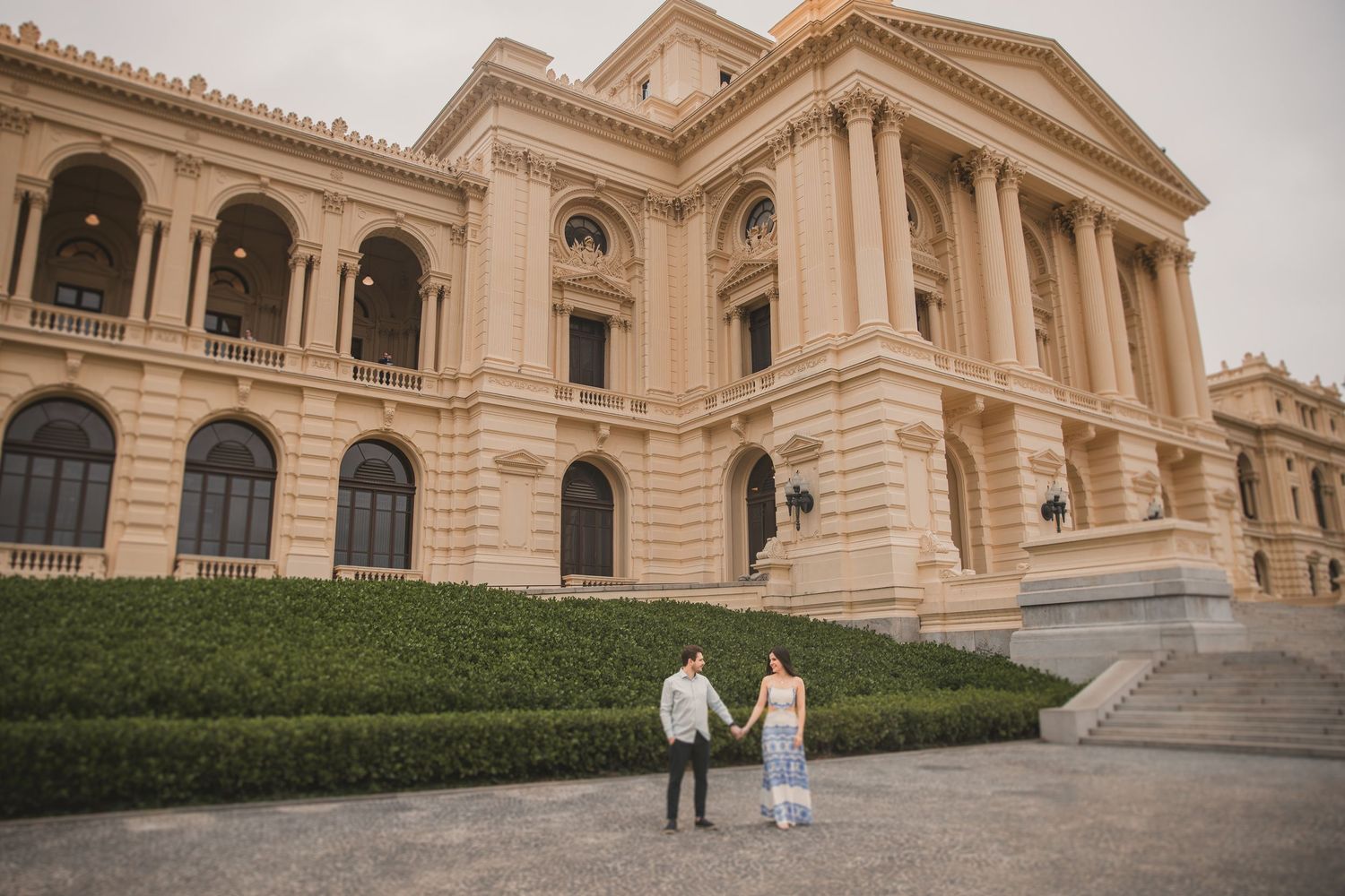 Two people stand in front of a grand neoclassical building with columned architecture and manicured hedges.