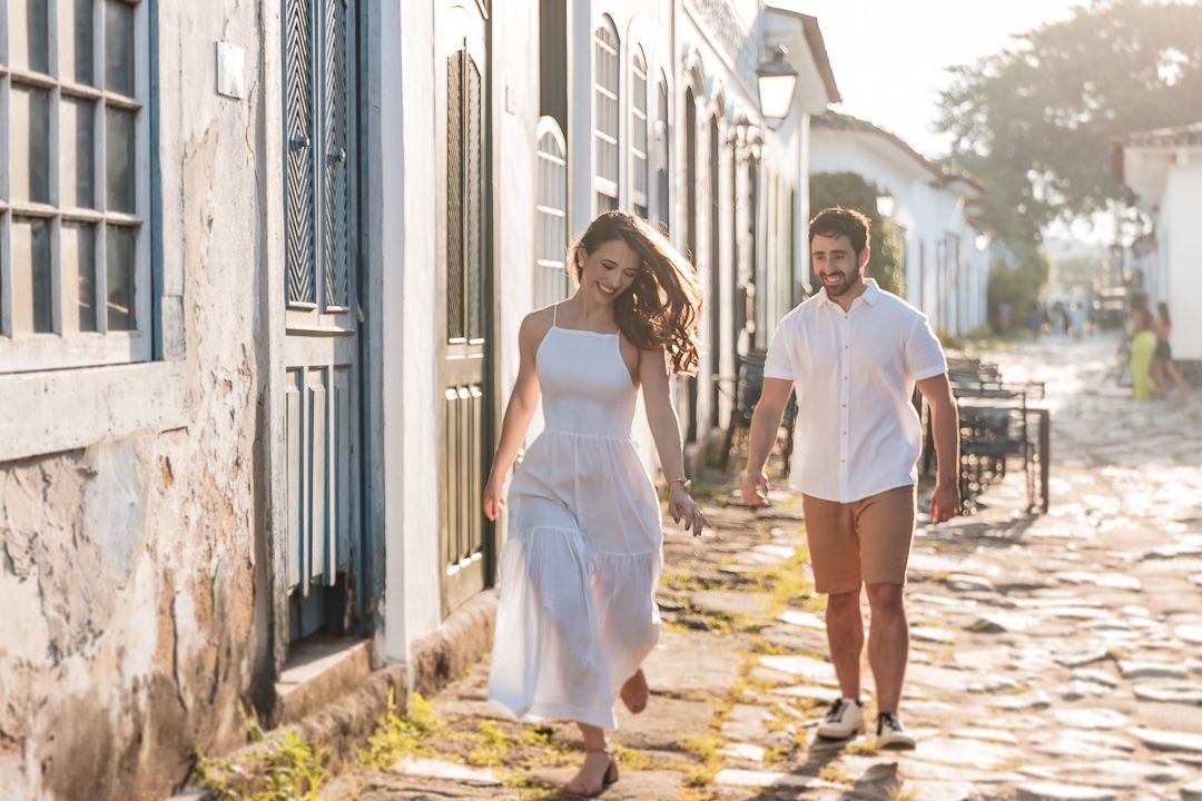 Couple strolling together on a charming cobblestone street during golden hour with white buildings in the background.