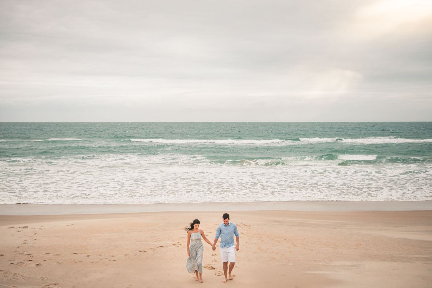 Two people walk hand in hand along a deserted beach with turquoise waves and cloudy sky in a romantic seaside scene.