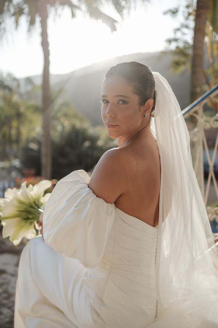 A bride in an off-shoulder white wedding dress and flowing veil poses outdoors with palm trees and mountains in the background.