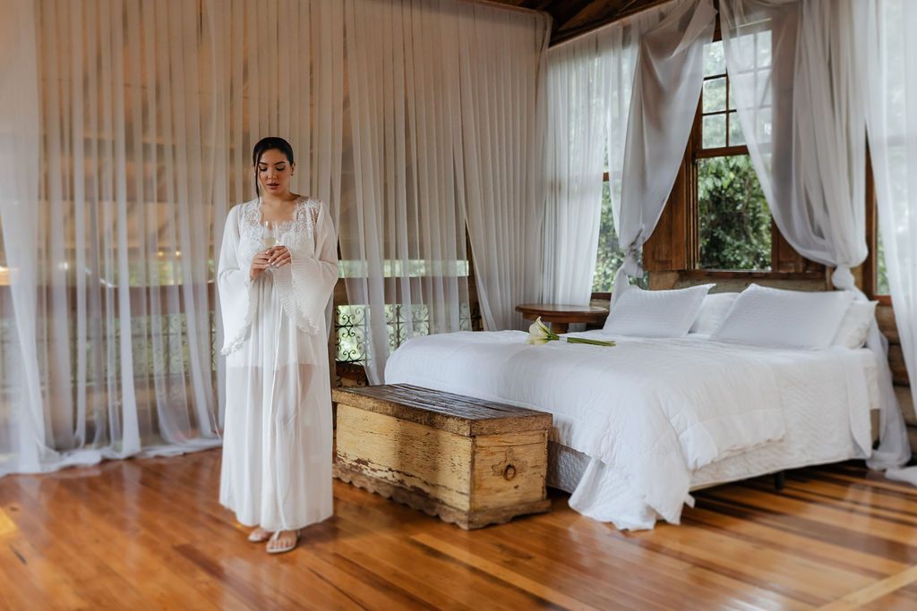 Fotógrafo de casaElegant vintage-style bedroom with wooden floors, sheer white curtains and four-poster bed featuring antique storage chest.