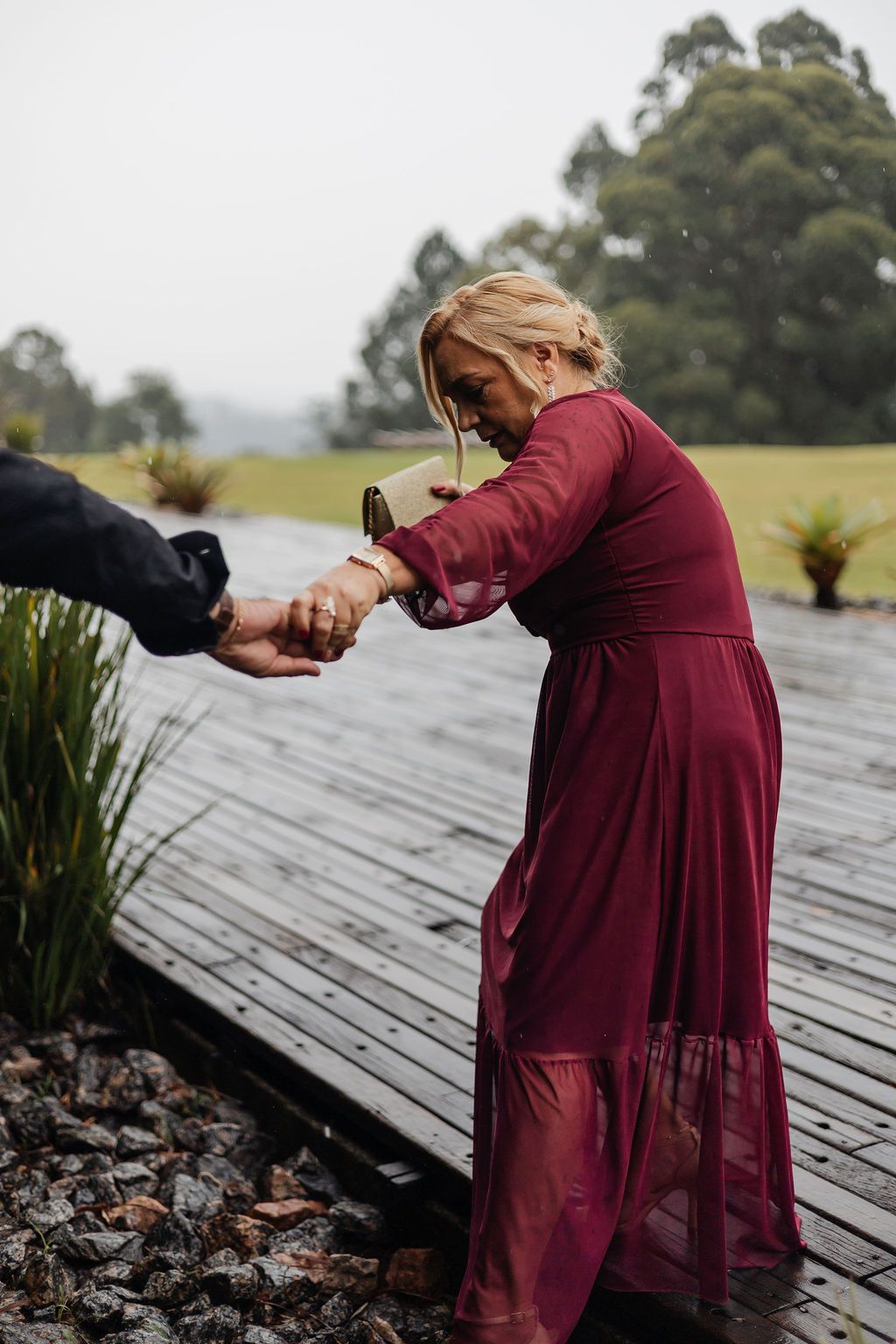 Inspire-se com o casamento ao ar livre em São PaA figure in a flowing burgundy dress stands on a wet wooden deck in a rainy outdoor setting.