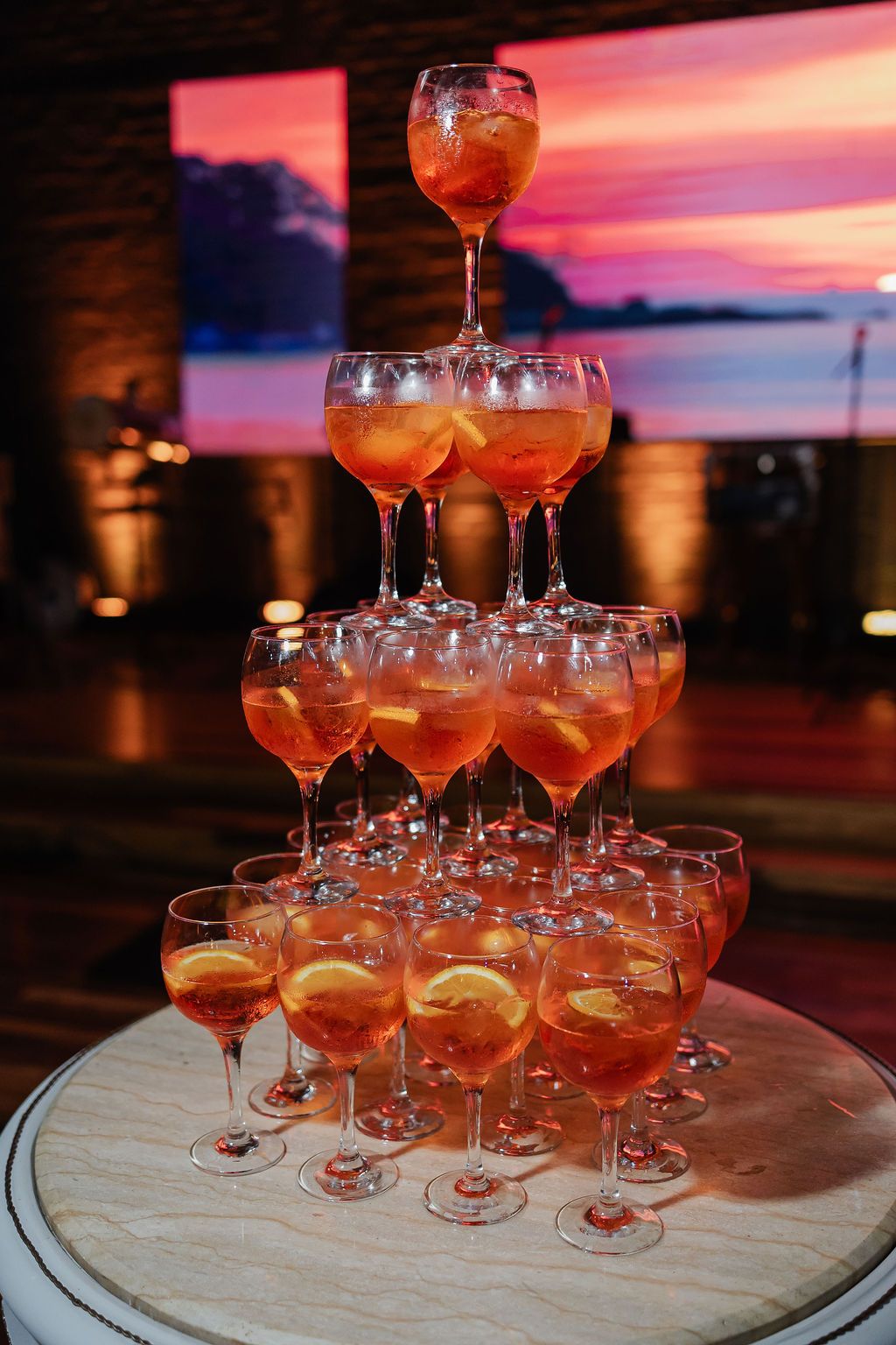 A tower of champagne glasses filled with orange-pink cocktails arranged on a round wooden surface against a sunset backdrop.