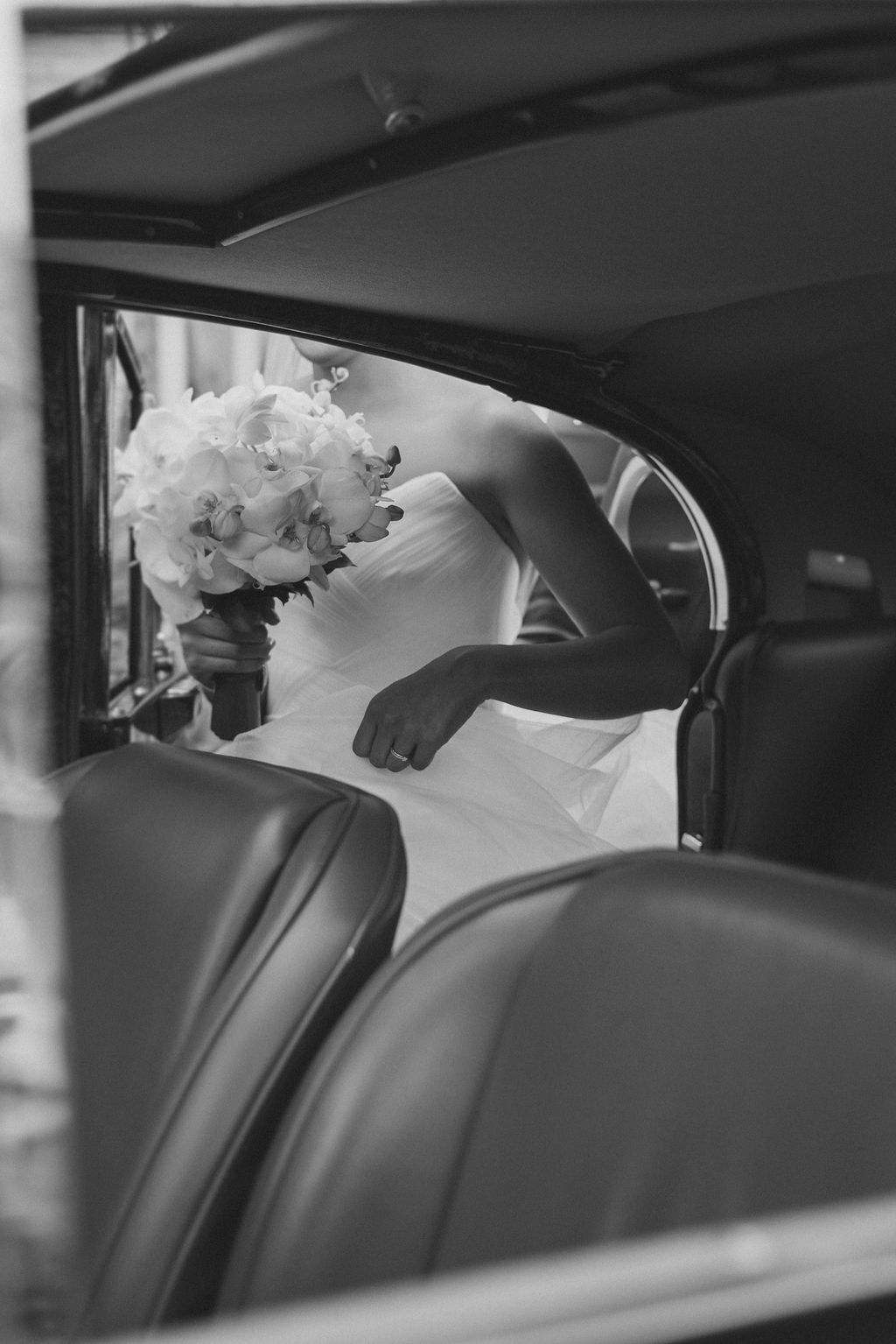 A black and white wedding photo showing a bouquet of white flowers as someone enters a vintage car interior.