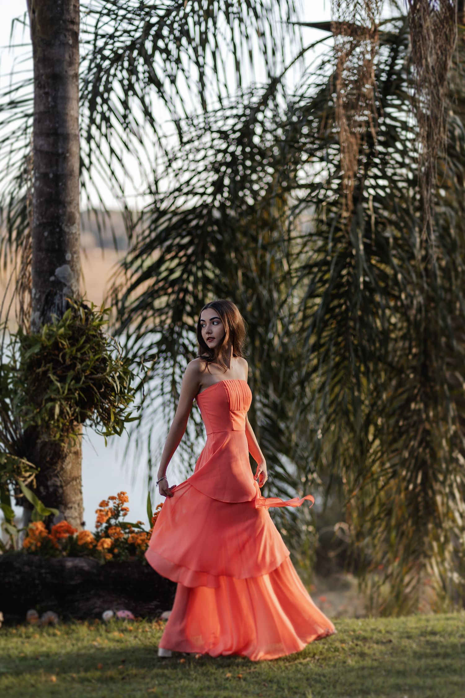 A figure in a flowing coral gown stands gracefully among palm trees in a tropical garden setting at sunset.