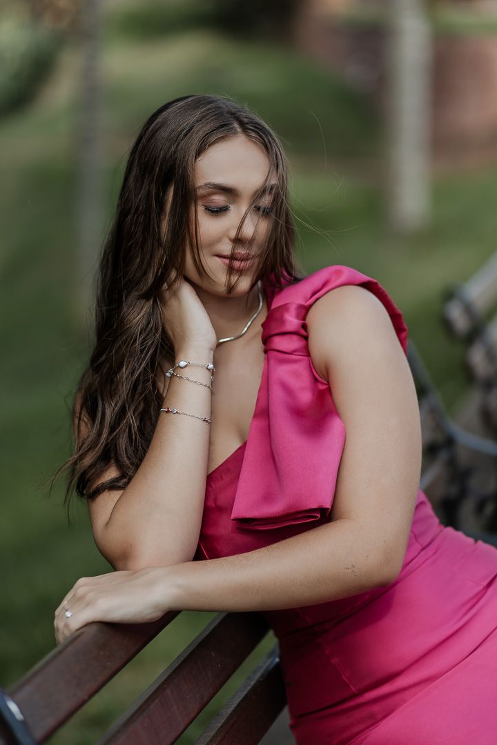 A woman in a vibrant pink dress poses thoughtfully while leaning on wooden railing outdoors.