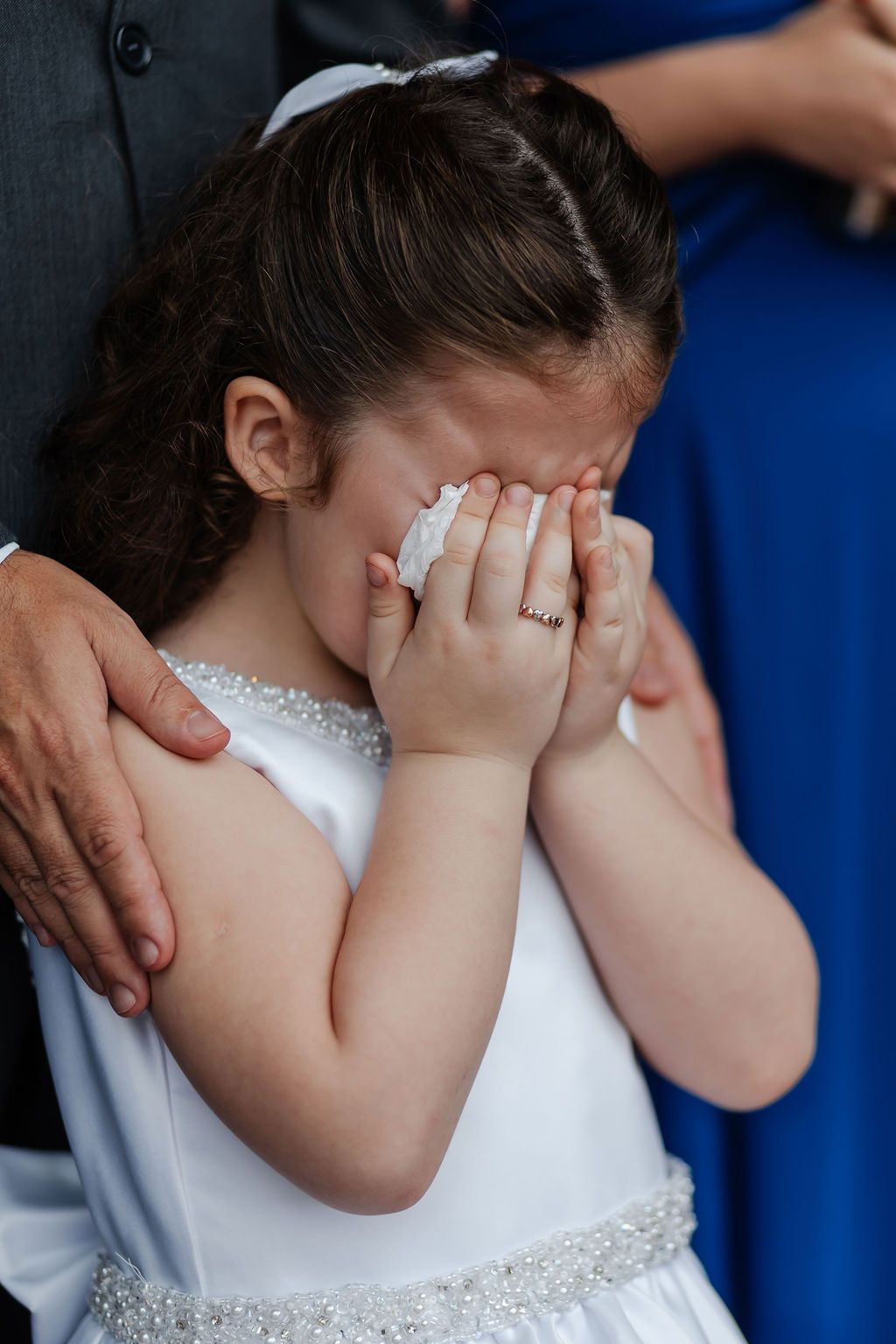 A small child in a white dress with sparkly details covers their face with both hands during an emotional moment.