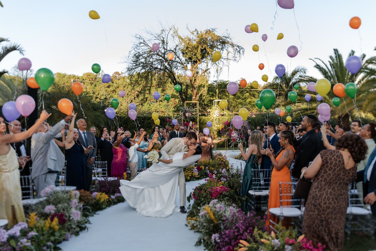 Colorful balloons float above an outdoor wedding celebration with vibrant floral arrangements and palm trees at sunset.