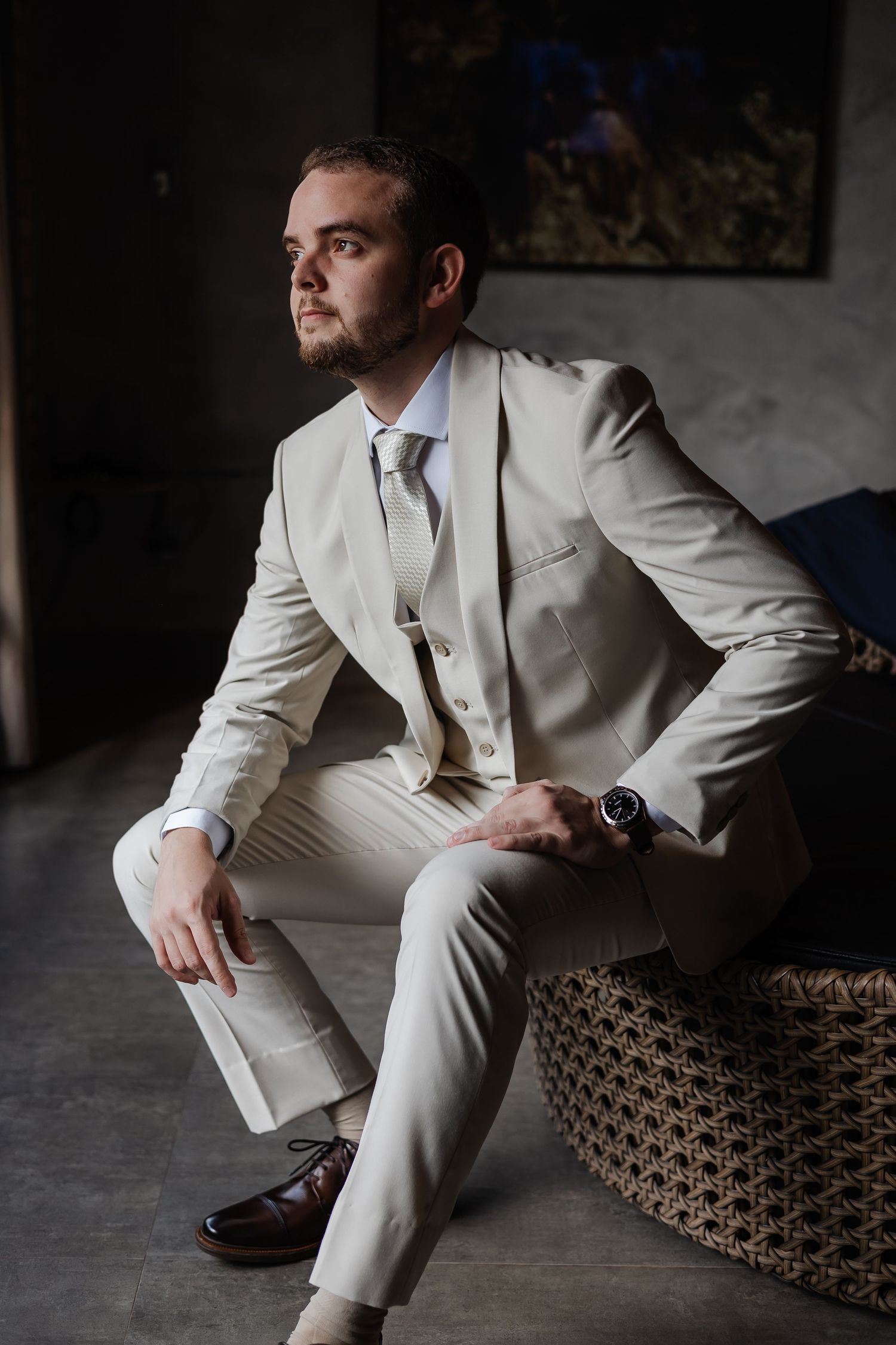 A person in a stylish beige three-piece suit with tie sits contemplatively on a woven ottoman in moody lighting.