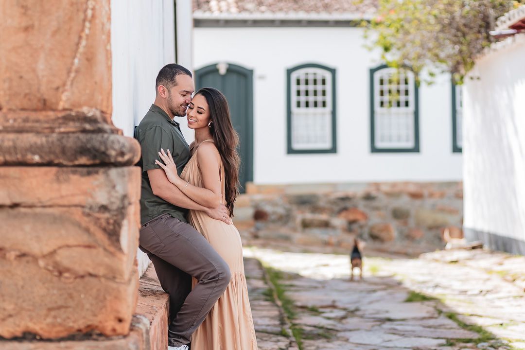 A romantic couple embraces against a rustic stone wall in front of a colonial-style building with arched windows.