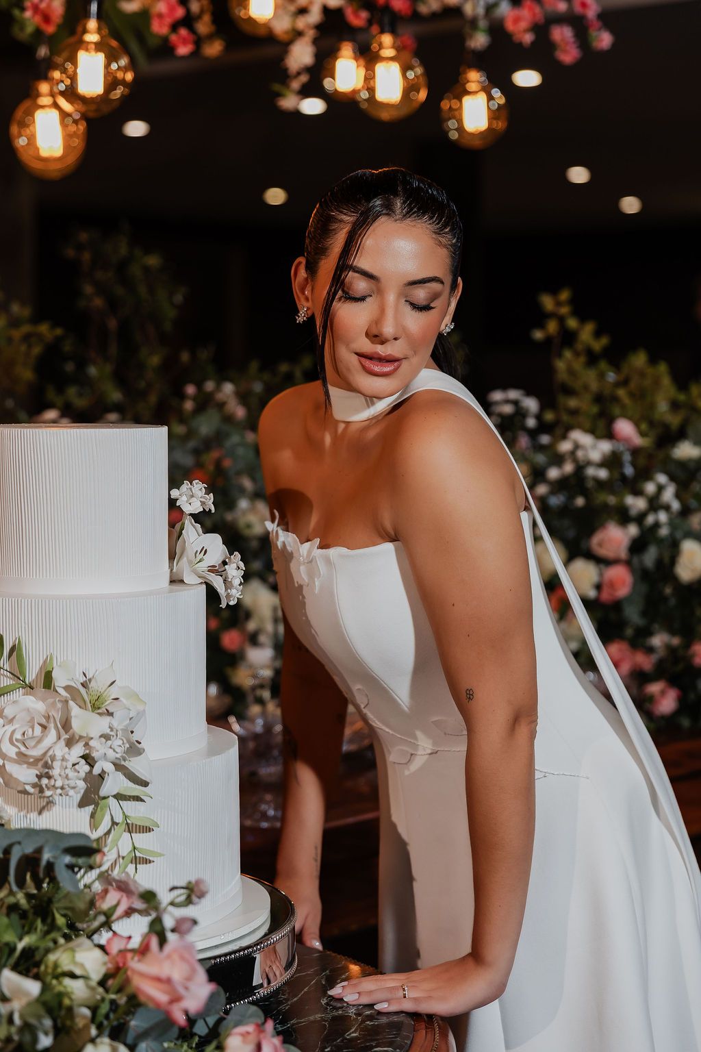 A candid moment of someone in a white wedding dress smiling while standing next to a floral-decorated tiered cake.
