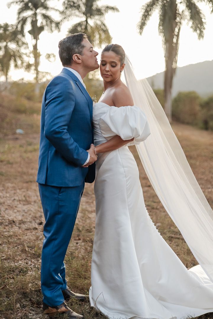 A romantic wedding photo of a couple in blue suit and white dress with flowing veil against a palm tree backdrop.