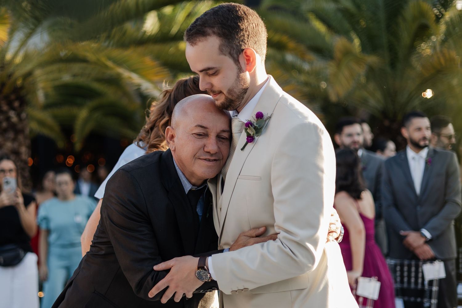 Two people sharing an emotional embrace during an outdoor wedding ceremony with palm trees and guests in the background.