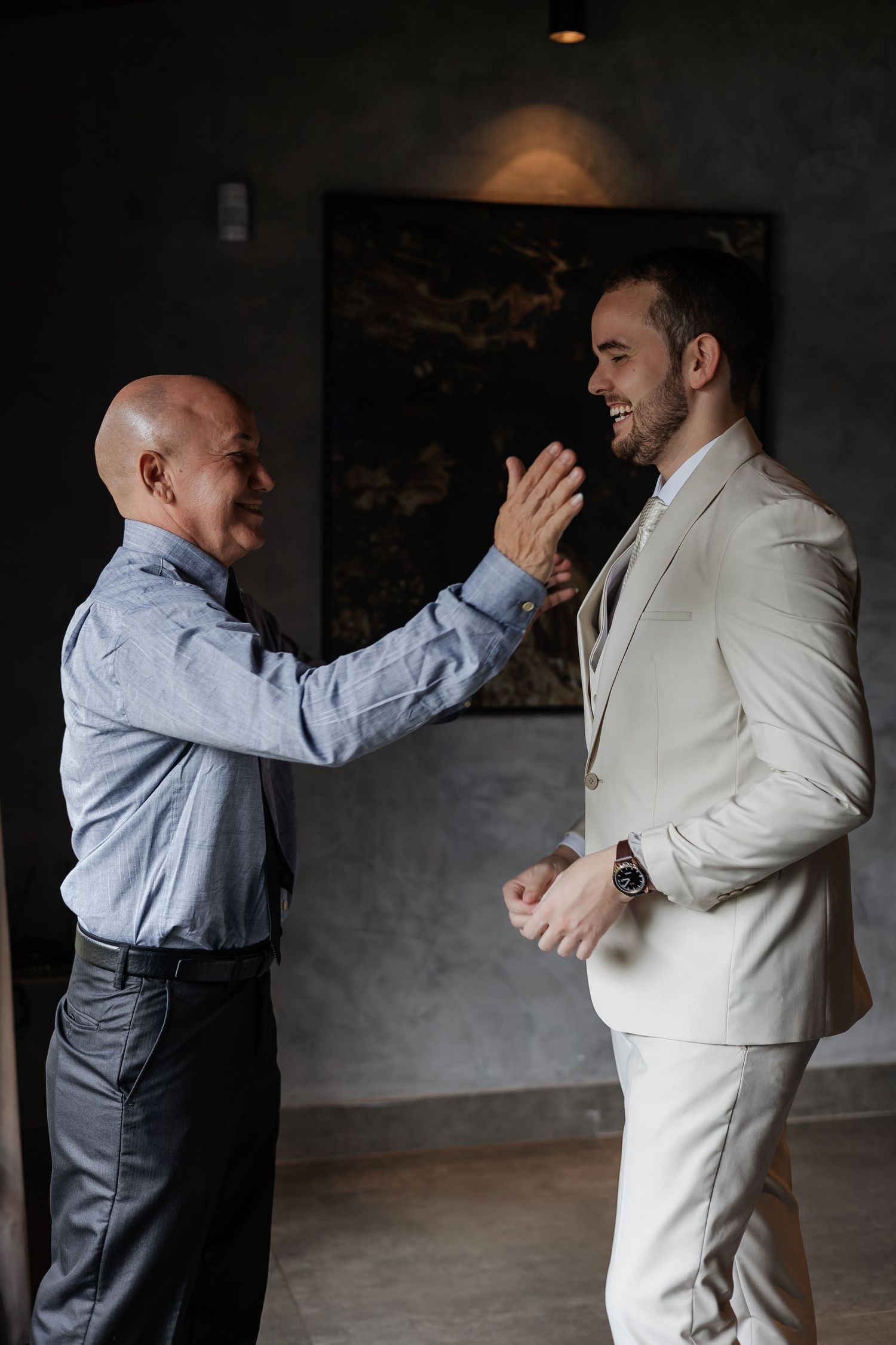 Two men share an emotional moment and laughter, one in a light suit and one in a button-down shirt in an indoor setting.